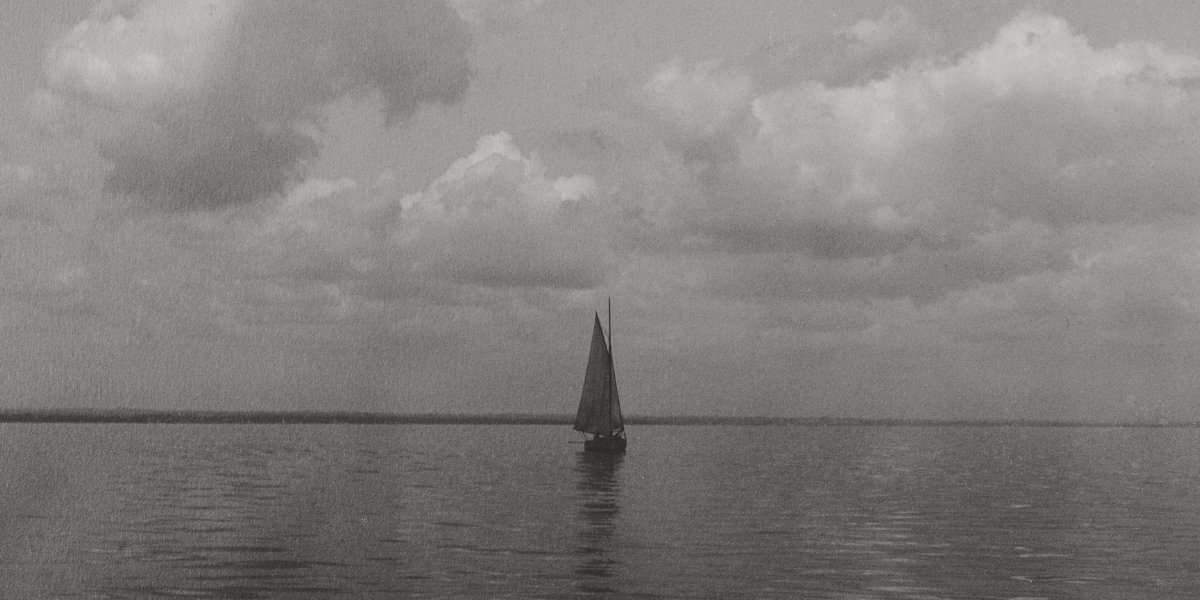 A monochrome photograph of classic sailboat gliding smoothly across vast, tranquil waters under a soft, overcast sky.
